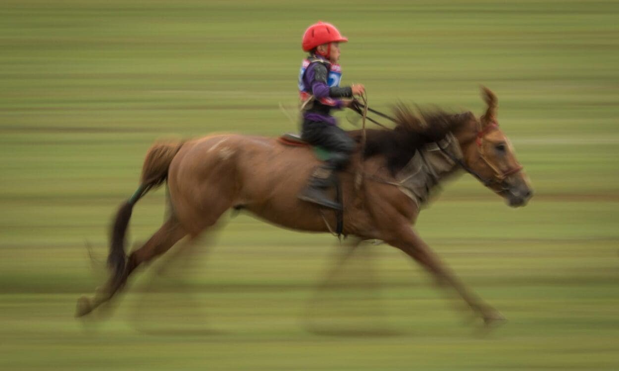 Young jockey in a red helmet racing horseback at a Naadam festival in Mongolia, background streaked with motion blur while horse and rider stay sharp