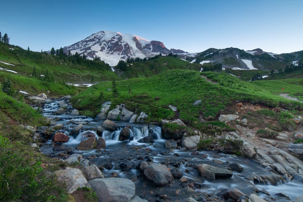 Small waterfall cascading over rocks in an alpine meadow at Mount Rainier, the peak glowing at sunrise in the background