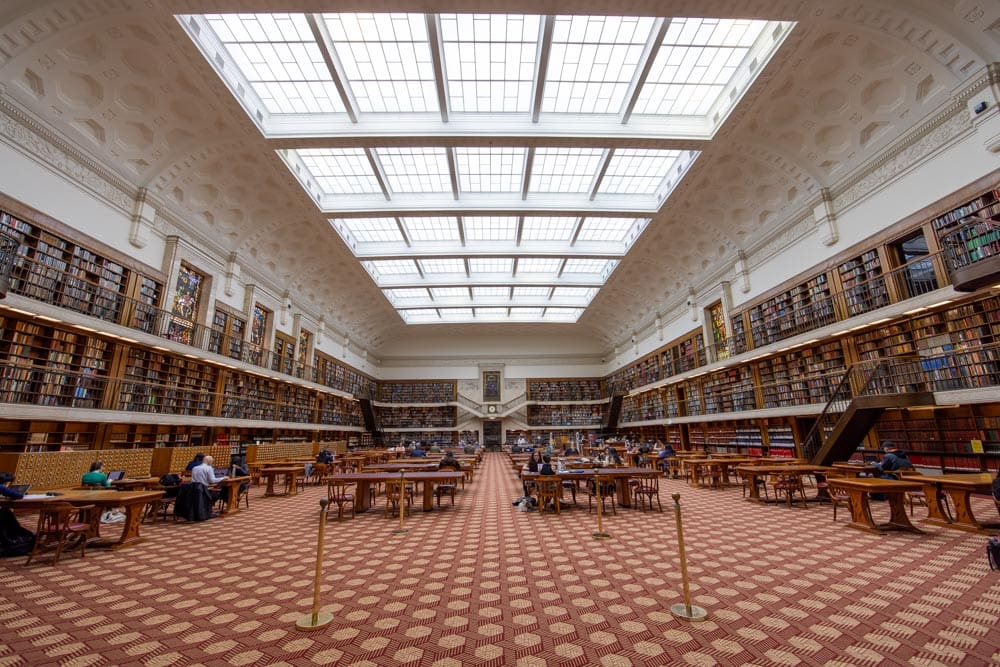 Grand reading room of the State Library of New South Wales in Sydney, Australia, with rows of bookshelves, wooden tables, and a large glass skylight overhead