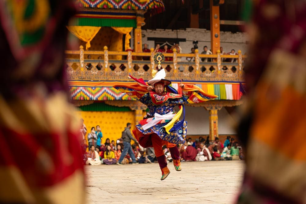 Costumed dancer leaping mid-air at the Paro Tshechu festival in Bhutan, elaborate patterned robe and hat, framed tightly between blurred foreground fabric