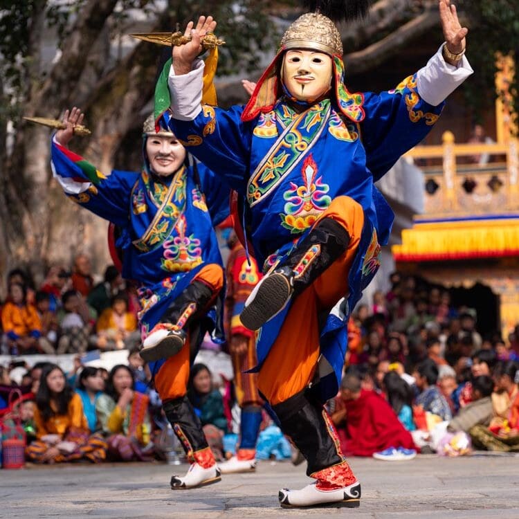 Two masked dancers performing at the Paro Tshechu festival in Bhutan, dressed in blue and orange traditional costume, ceremonial daggers raised, seated crowd in the background