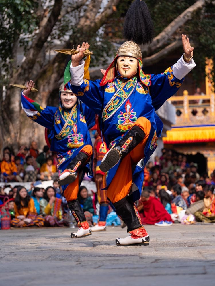 Two masked dancers performing at the Paro Tshechu festival in Bhutan, dressed in blue and orange traditional costume, ceremonial daggers raised, seated crowd in the background