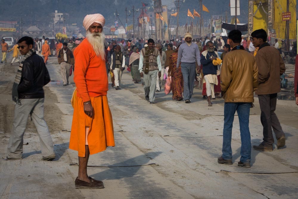 Man in an orange robe and white turban standing still on a crowded street in Varanasi, India, while pedestrians move around him