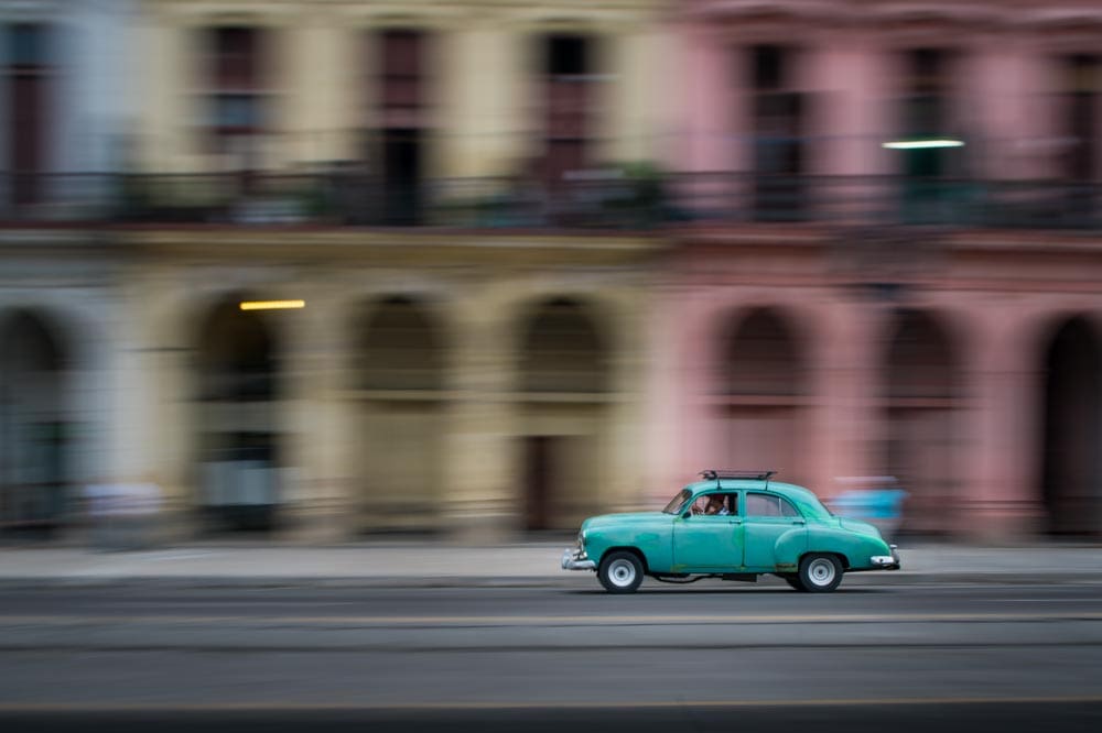 Teal vintage car panned along a street in Havana, Cuba, colorful building facades streaked into horizontal blur