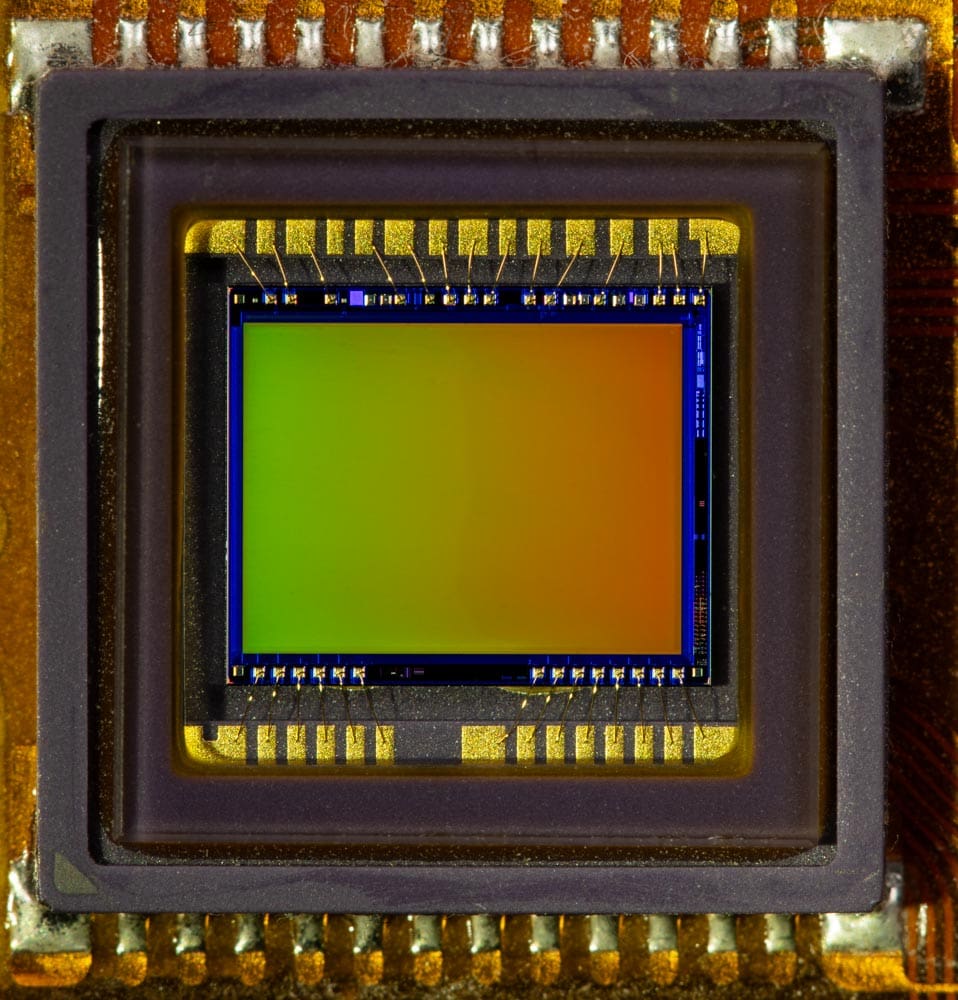 Macro close-up of a camera image sensor chip, showing its colorful surface and wire-bonded connections.