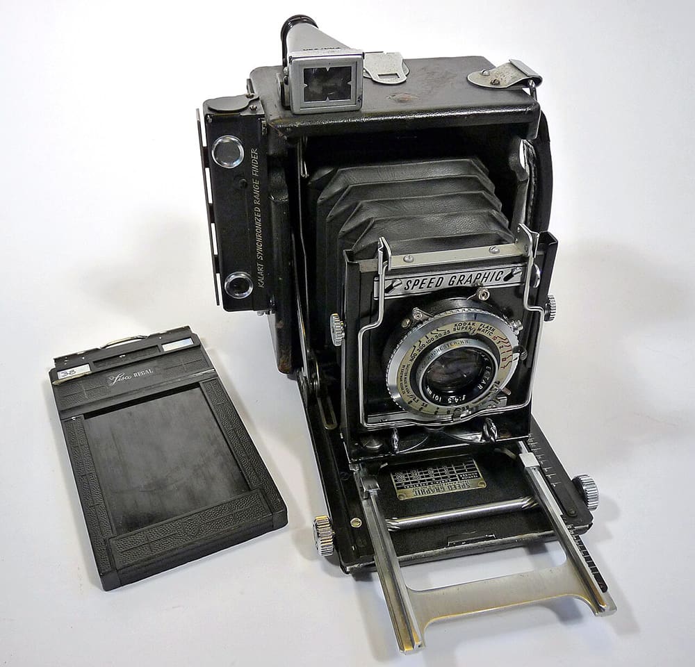 A vintage black Speed Graphic press camera with bellows, next to a film holder.