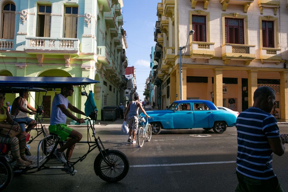 A busy Havana street scene with a bright teal vintage car, a bicycle taxi, cyclists, and pedestrians framed between pastel-colored colonial buildings.