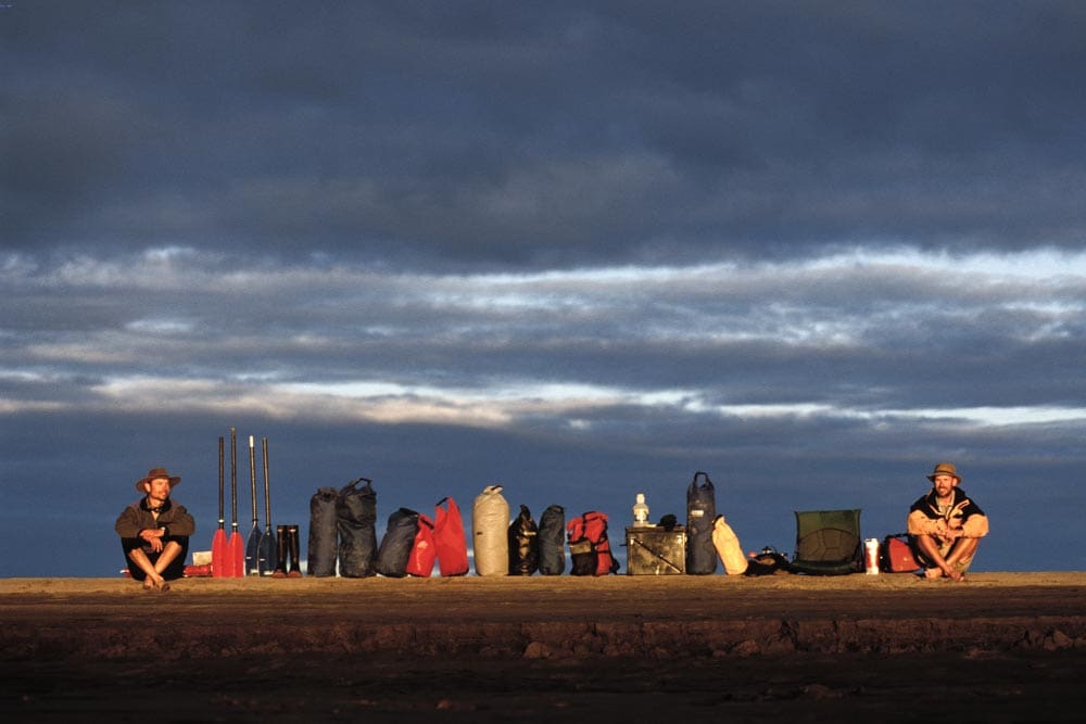 Two men sit at opposite ends of a long row of camping and canoeing gear lined up on a muddy riverbank at dusk, under a dramatic layered sky.