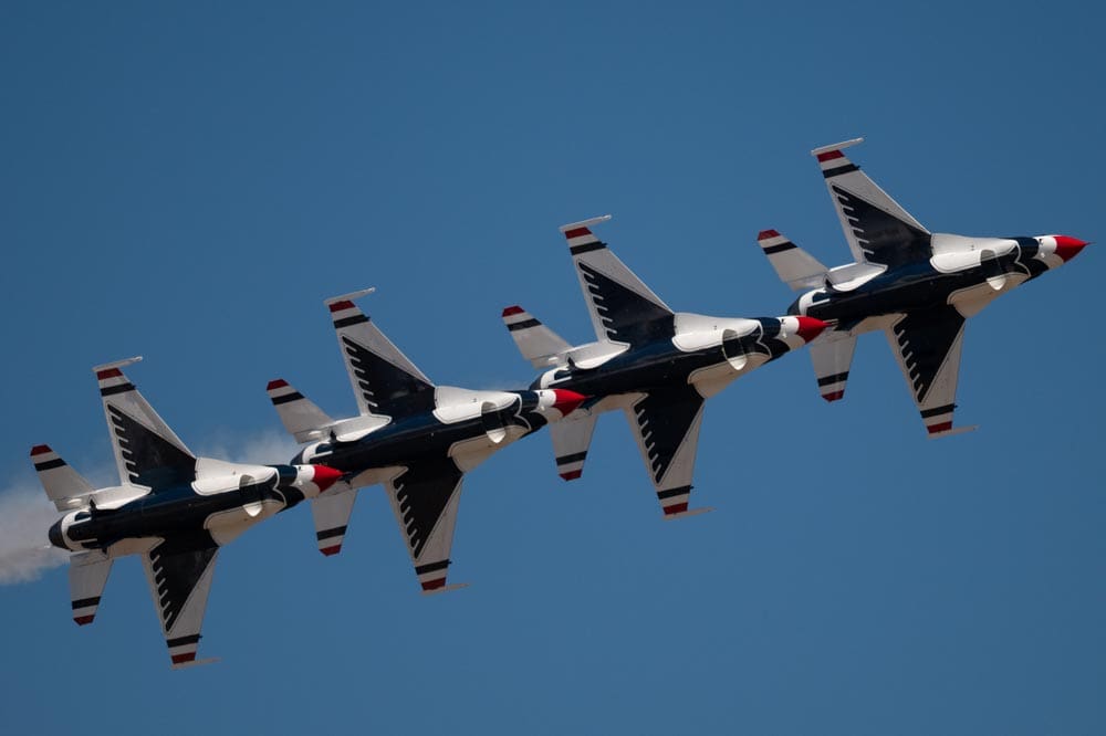 Four Air Force Thunderbird jets flying in tight diagonal formation against a clear blue sky, trailing a thin line of smoke.