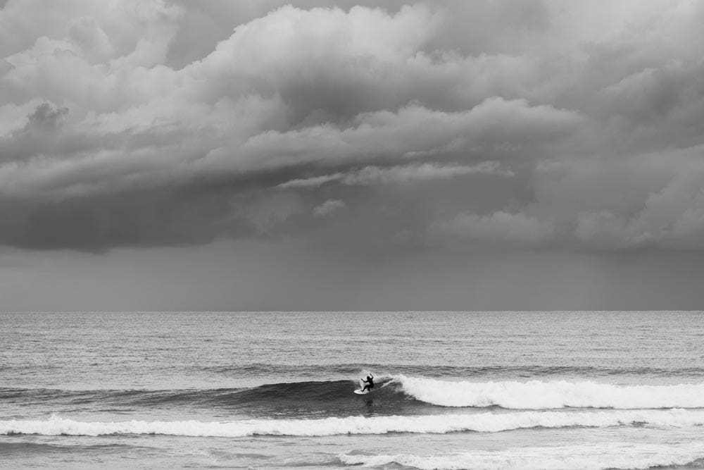 Black-and-white photo of a lone surfer riding a small wave, dwarfed beneath a massive bank of storm clouds over the ocean.