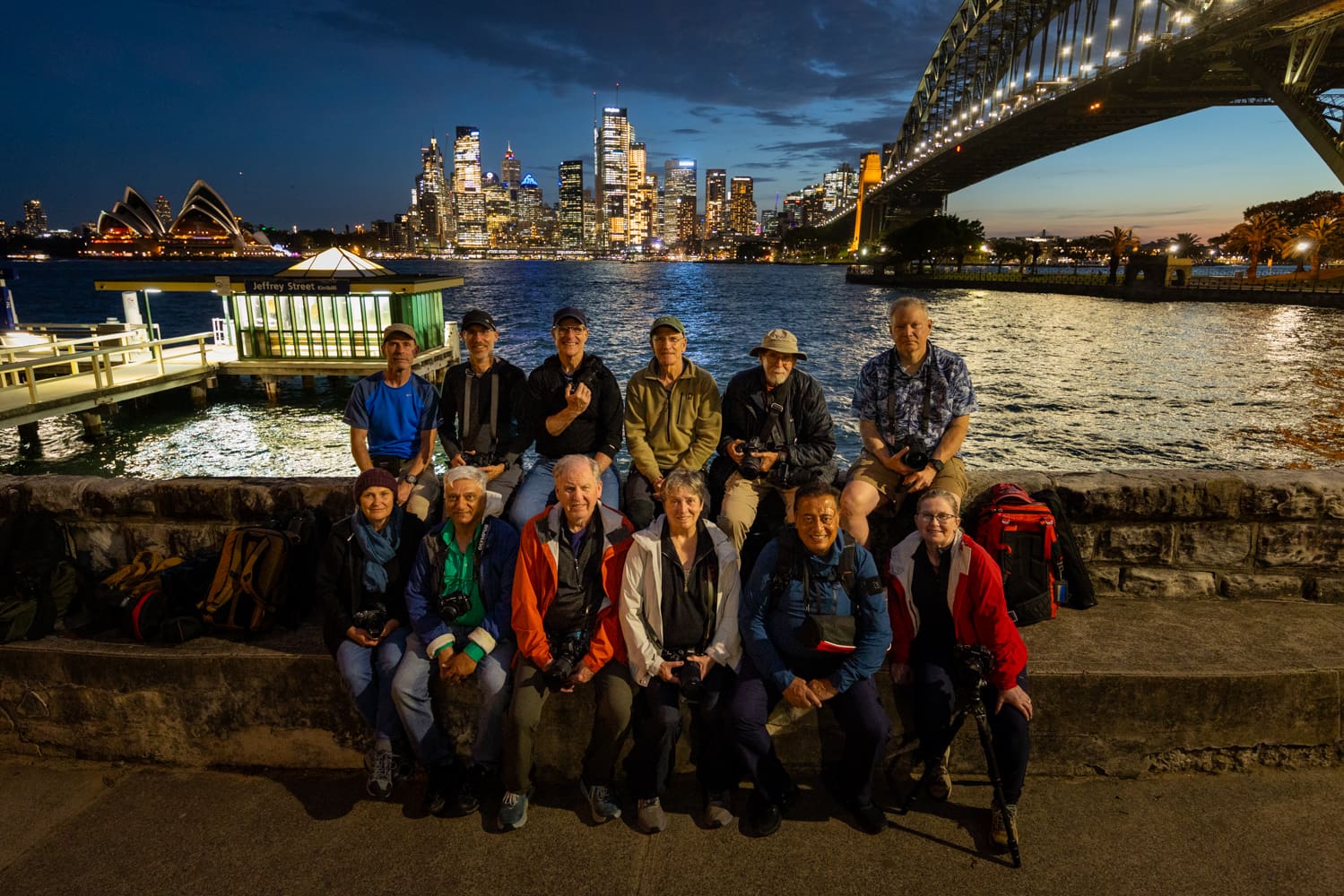 Ten tour photographers with with tour leaders pose at night by Jeffrey Street Wharf, with the Sydney skyline and Harbour Bridge behind them