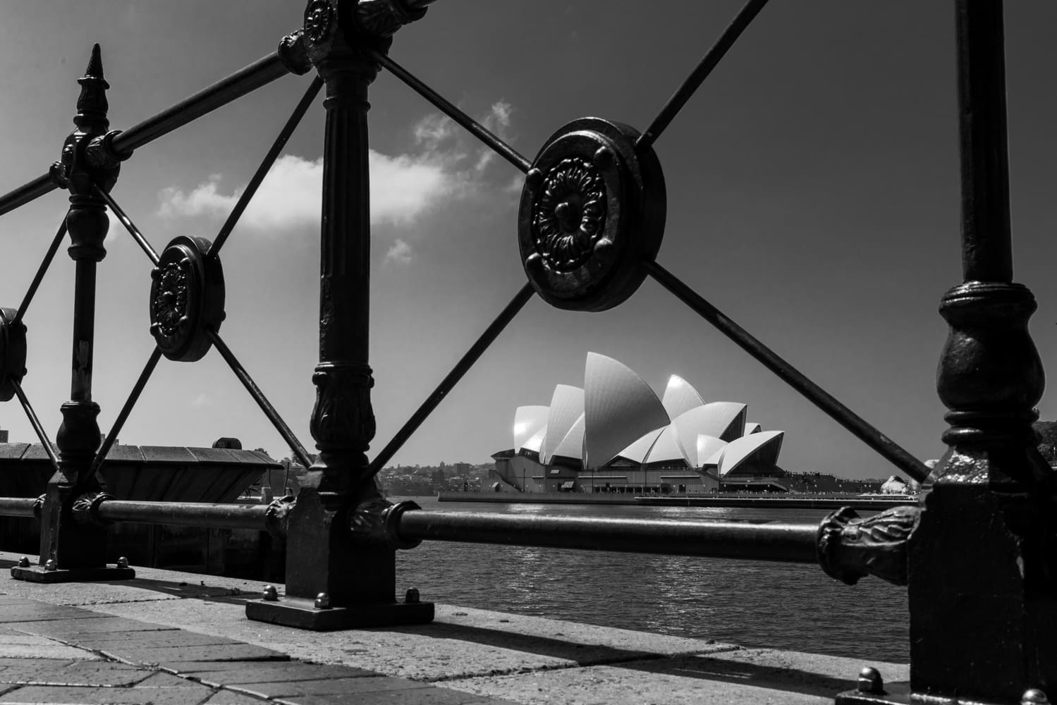 Black-and-white view of the Sydney Opera House framed by an ornate iron railing across the harbor