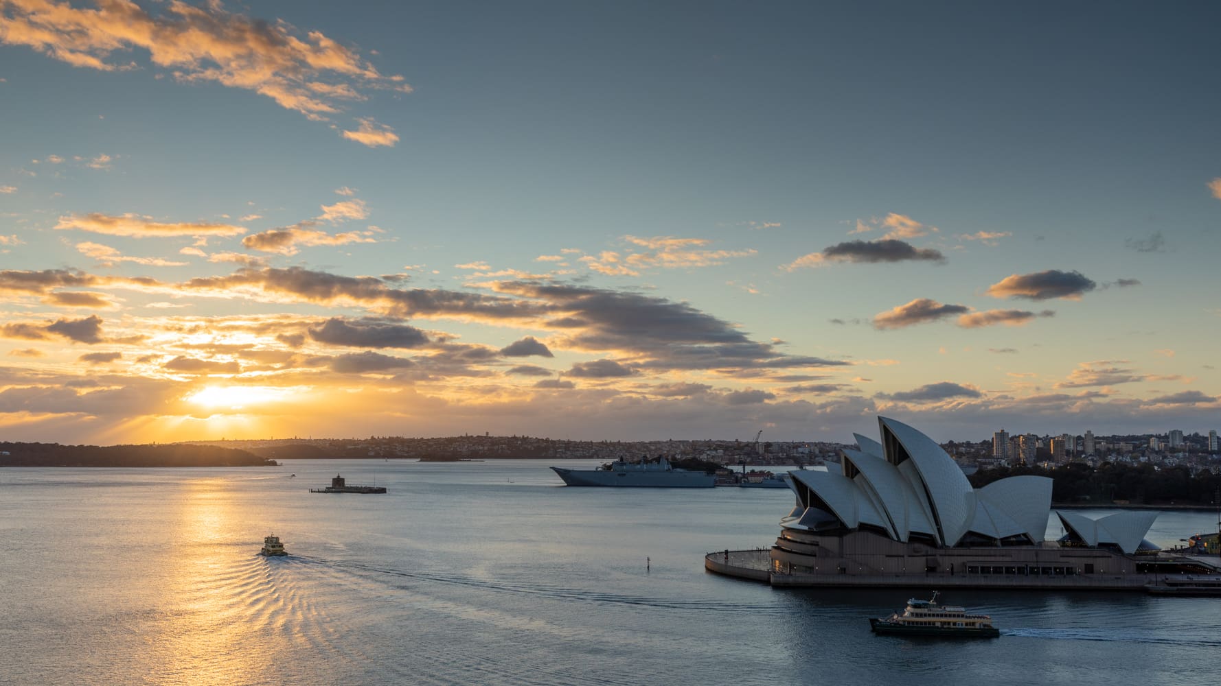 Sunrise breaks through clouds over Sydney Harbour, with the Opera House and ferries below, seen from the Harbour Bridge