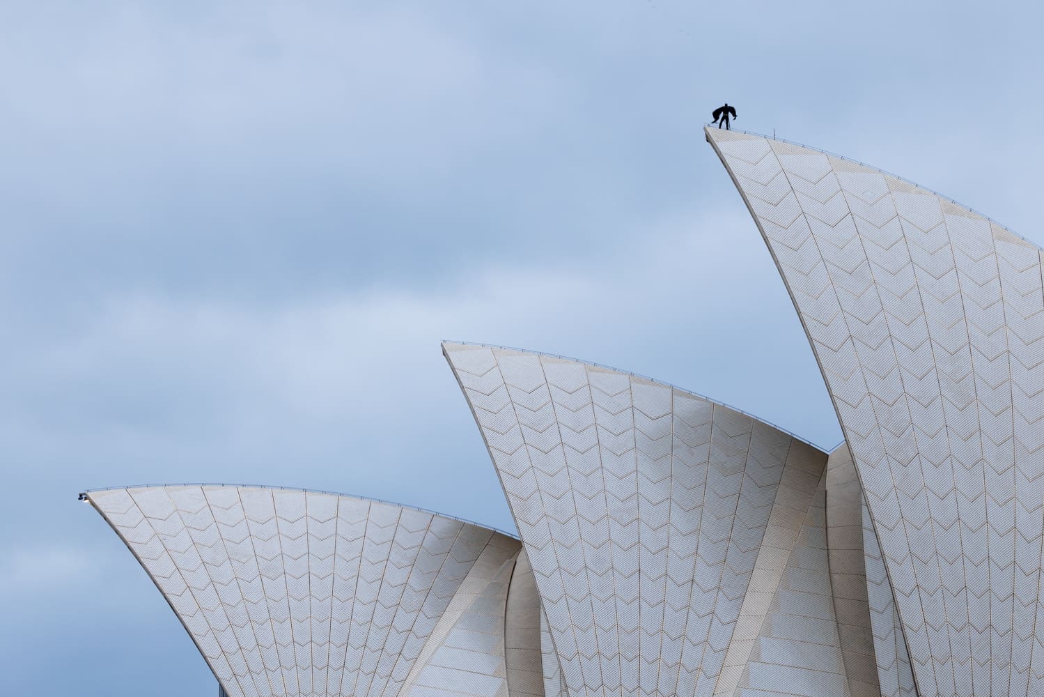 A person dressed as Batman stands on top of the highest sail of the Sydney Opera House against a cloudy sky