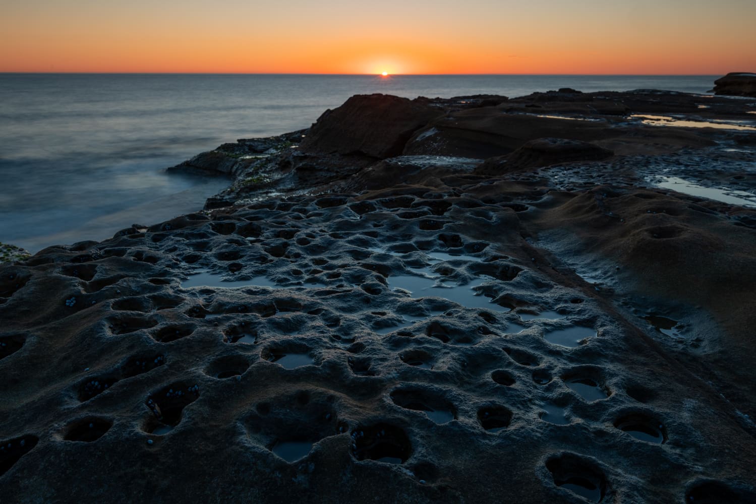 The sun rises over the ocean beyond a sandstone rock shelf pitted with small tide pools