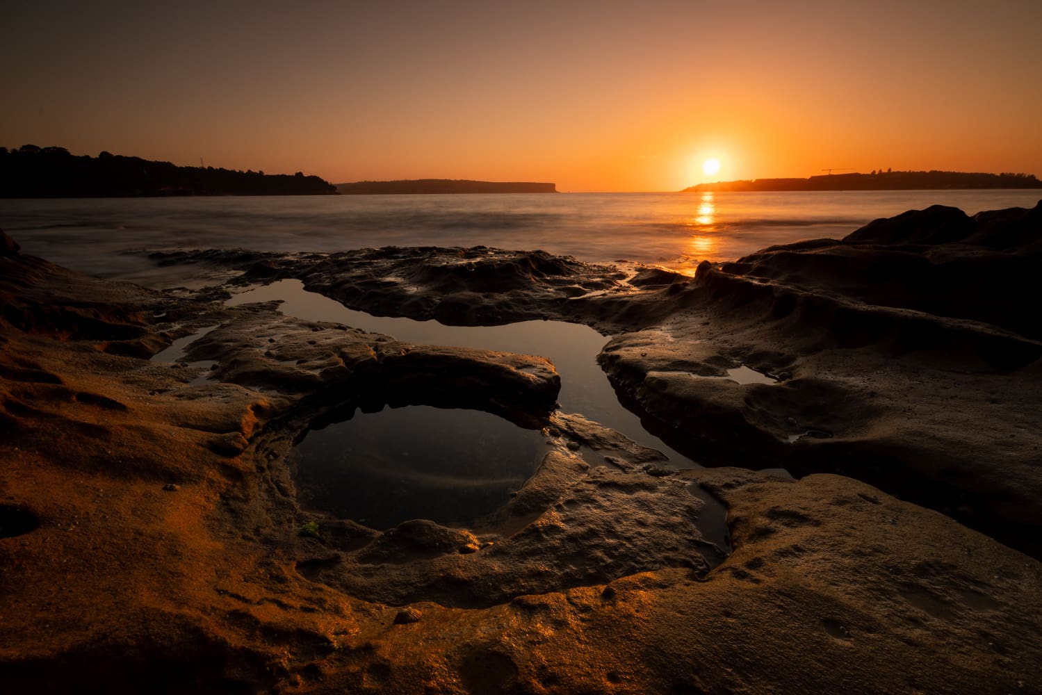The sun rises between the Sydney Heads, seen across tide pools on a sandstone rock shelf at Middle Head